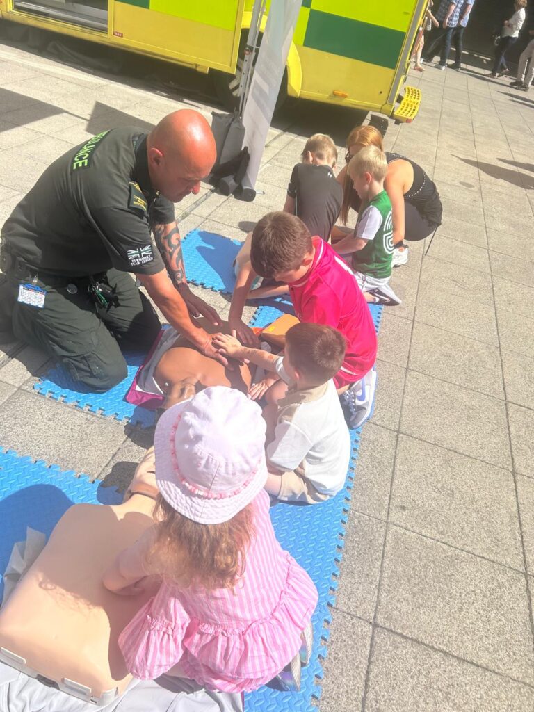 The image shows children being taught how to restart a heart by a member of EEAST.